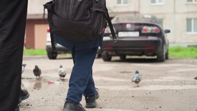 Child walking with backpack and parent on urban pavement, pigeons nearby, parked cars, boots splashing small puddles, jeans and jacket, steady stride preparing for school commute, morning routine,