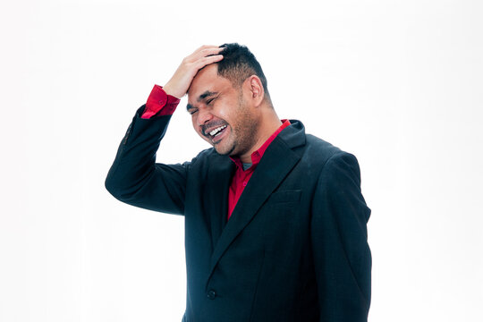 Portrait of a joyful man in a black suit and red shirt laughing while touching his forehead in a facepalm gesture, isolated on a white background, representing a funny mistake, "oops" moment, or light