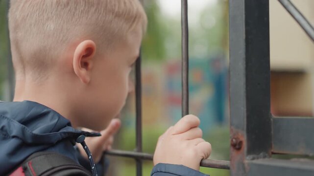 Young boy at school gate ready with backpack, navy jacket and steady grip on metal bars, hopeful gaze into campus, preparing to go back after break, clear daylight, closeup hands and profile, composed