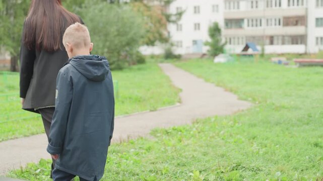 Caucasian boy and guardian walking to school overcast suburban playground path, mother carrying bag, boy looking down, hesitant steps, jacket hood, quiet morning, prepare for school day, emotional
