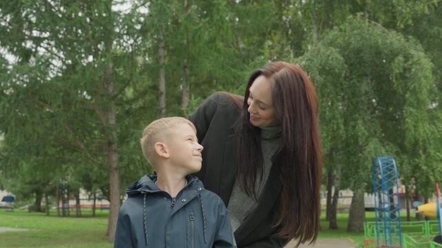 Mother and white boy reassuring kiss, warm sendoff in park with affectionate gesture and confident smile, last minute comfort and encouragement before leaving for school, leafy playground background