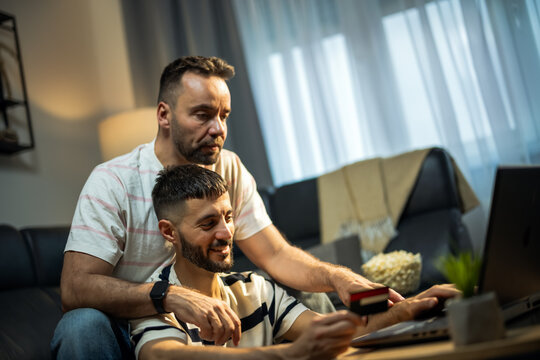 A happy same sex male couple using a laptop and credit card to shop online in their living room.