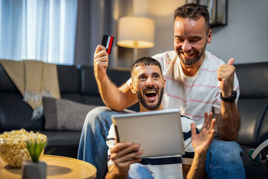 Triumphant same sex couple raising hands after confirming an online order on a laptop at home.