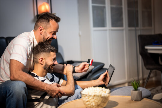 A happy same sex couple holding a credit card and shopping with a digital tablet on a sofa.