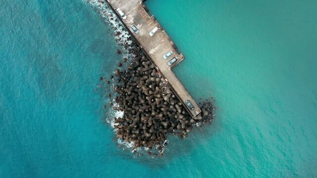 4k Aerial shot panning up to reveal a concrete pier with parked cars and defensive tetrapods extending into clear blue ocean water
