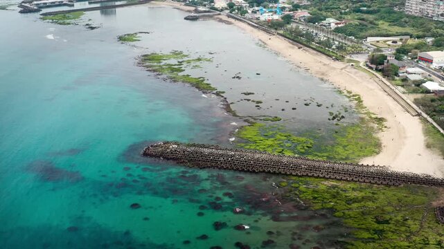 Aerial high angle view of a long concrete breakwater extending from a sandy beach with a green algae covered reef into clear turquoise ocean water