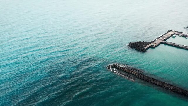 4k Aerial high angle view of a coastal marina entrance featuring a long concrete breakwater and tetrapods extending into vast calm blue ocean water
