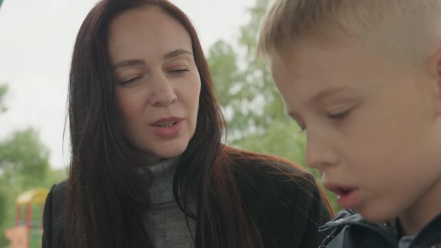 Mother and son prepare backpack outdoors on park bench, cloudy morning, mother gently guides son while he packs jacket and books, intimate supportive moment showing parental care and school routine