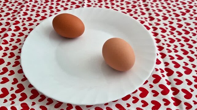 Two brown eggs rest on a white plate atop a tablecloth patterned with repeating red hearts, creating a simple yet striking contrast between everyday food and vibrant background