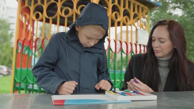 Boy concentrating on school supplies, arranging pencils and paper under adult supervision at outdoor bench, markers and workbook visible, handson preparation, rainy hood, diligent expression