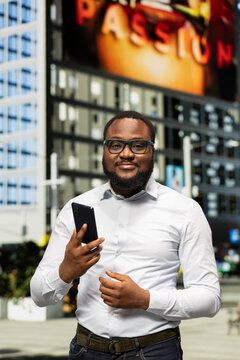 Trendy black adult browsing on his smartphone through a downtown avenue, walking around vibrant buildings and expressing a confident attitude. Independence in modern metropolis.