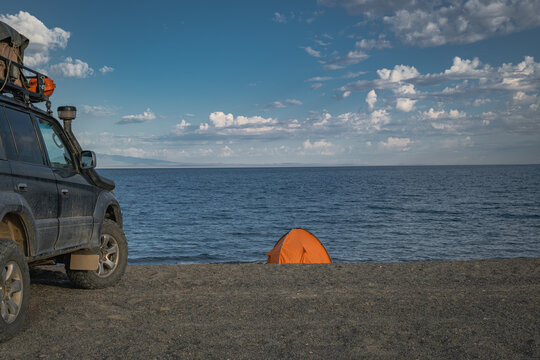 Off-road 4x4 car with expedition roof rack parked on a lake shore near orange tent. Camping and overlanding lifestyle concept in wild nature under a cloudy sky.