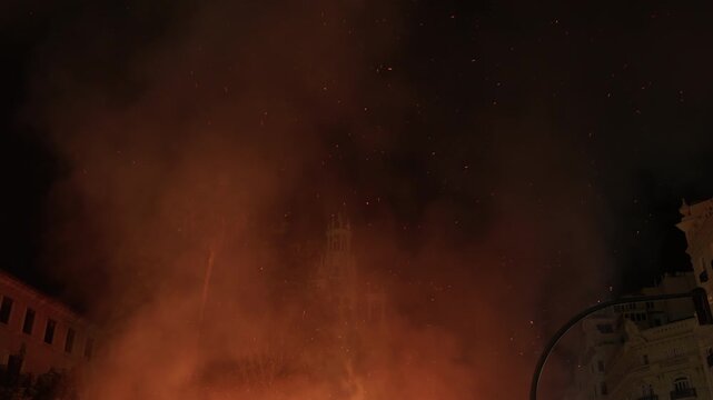 Abstract upward view of embers and orange smoke rising from a burning falla into the dark sky above city buildings during Las Fallas