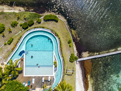 Aerial view of a luxury resort swimming pool with people swimming next to a wooden deck and a pier extending into the turquoise ocean in Guanaja, Bay Islands Department, Honduras.