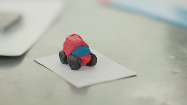 Clay toy car on desk card bright red and blue handmade model rests on paper card next to notebook and smartphone playful classroom atmosphere soft daylight shallow focus student preparing back