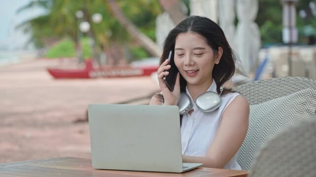 Cheerful young Asian woman enjoys remote work at a seaside cafe, talking on her smartphone and using a laptop with headphones around her neck during a tropical vacation.