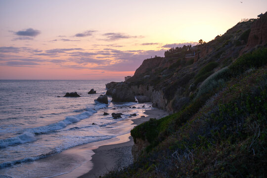 Sweeping coastal overlook at sunset showing pink-lavender sky, scattered sea stacks, sandy cove, eroded sandstone cliffs, and park staircase at El Matador State Beach, Malibu CA. California travel and