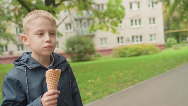 Park path, white boy walking with cone preparing to head back to school. Boy licks melting ice cream, thoughtful expression, jacket up, residential buildings and green lawn in background, quiet