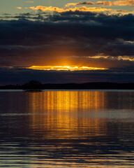 Fototapeta premium Dramatic Golden Sunset Breaking Through Dark Storm Clouds Over Finnish Lake with Island Silhouette