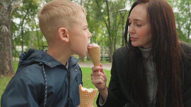 Mother guides son. Caring mom helps young boy. Gentle teaching in park setting. Warm interaction as mother offers cone to child. Parent imparts guidance to son during playful moment on park bench