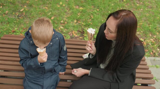 Caucasian mother and schoolboy sharing ice cream on park bench preparing for backtoschool, mother offers calming pep talk while schoolboy in navy jacket eats cone, autumn leaves scattered on ground,