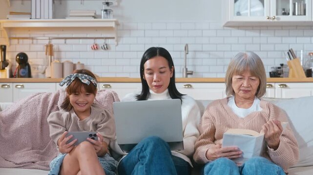 Asian three generations family enjoying time together on a sofa. The grandmother is reading a book, the mother is working on a tablet, and the young girl is playing on a smartphone at home