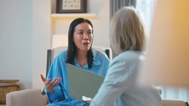 Senior Asian female psychologist taking notes during therapy session with woman patient.
