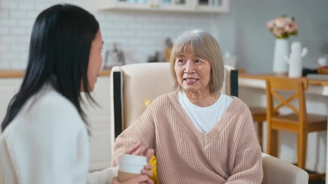 Asian adult daughter listens attentively while holding hands with her elderly mother over coffee. They are sharing a moment of emotional support in a cozy, warm living room at home together.