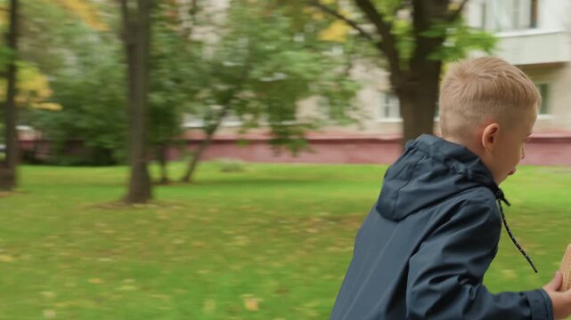 Young boy preparing for school, walking along leafy park path with backpack on bench, eating snack, wearing dark jacket, focused expression, slow deliberate pace, weekday autumn morning