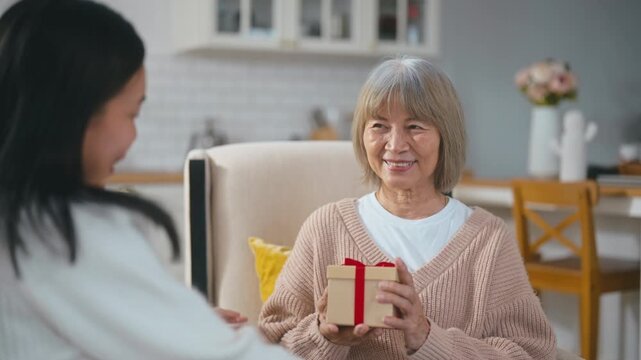 Daughter giving a gift to her elderly mother, who smiles happily and hugs her daughter on the living room sofa, showing love, family bonding, and warmth in a cozy home setting.