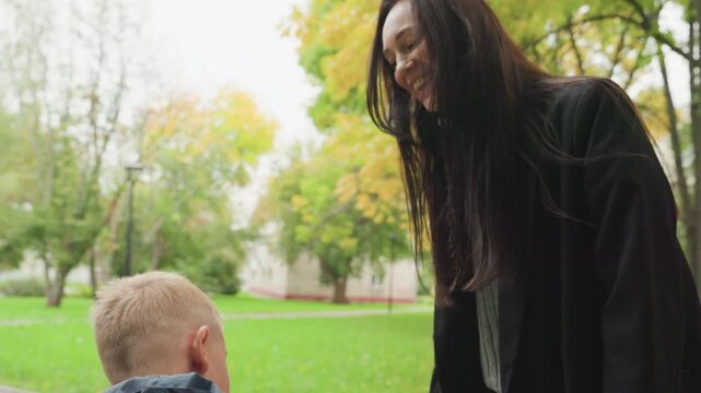 Woman prepares boy back to school, smiling caregiver gently adjusts jacket and hands small item to child in leafy park during autumn morning, warm farewell and supportive guidance, candid interaction