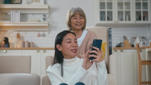 Elderly Asian mother is lovingly tying her adult daughter's hair in a cozy living room. The daughter is playing on her mobile phone and showing the screen to her mother, sharing a happy moment