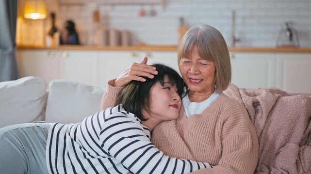 Asian adult daughter leans on her elderly mother's chest as the mother gently strokes her hair. Show bonding and love in a cozy, warm living room at home