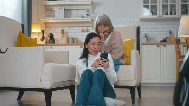 Elderly Asian mother is lovingly tying her adult daughter's hair in a cozy living room. The daughter is playing on her mobile phone and showing the screen to her mother, sharing a happy moment