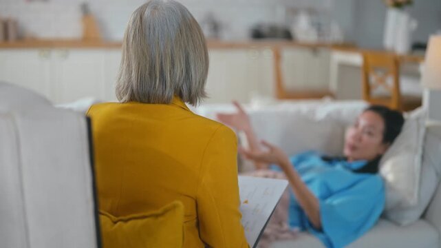 Senior Asian female psychologist attentively taking notes while listening to an Asian woman patient lying on a sofa during a therapy session, providing mental health support and counseling.