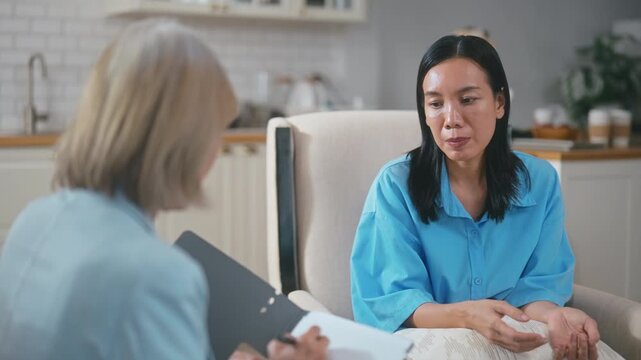 Senior Asian female psychologist taking notes during therapy session with woman patient.