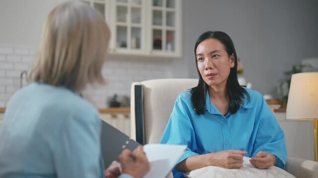 Senior Asian female psychologist taking notes during therapy session with woman patient.