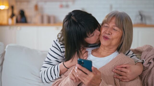 Slow motion Senior mother and daughter sitting on the sofa at home, looking at a smartphone screen together. Watching social media, shopping online, browsing news, and viewing pictures on the device.