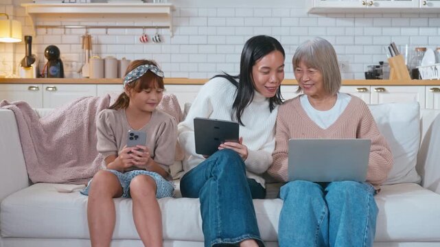 Slow motion Asian three generations family sits together on a sofa. The young girl is playing on a mobile phone, the mother is working on a tablet, and the grandmother is using a laptop in a cozy, mod
