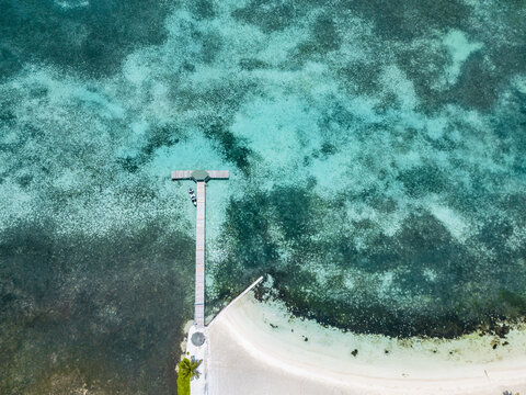Aerial view of a wooden pier extending into turquoise tropical waters with coral reefs and a white sandy beach in Guanaja, Bay Islands Department, Honduras.