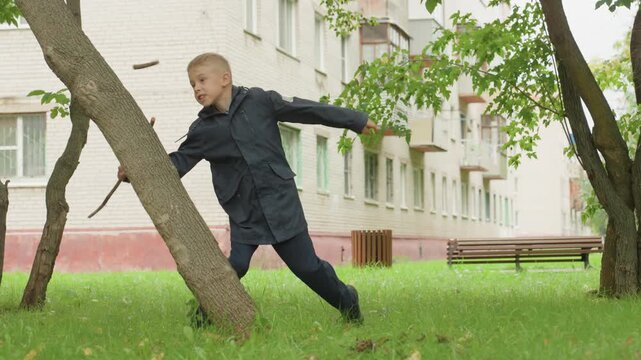 White schoolboy practicing stick combat moves in grassy courtyard near apartment block wearing dark jacket energetic jumps and lunges pretending swordplay playful training before back to school lively
