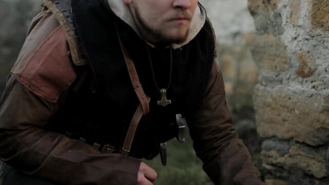 Mid close-up of medieval warrior kneeling and smelling soil held in fist against ancient stone ruins background. Cinematic tracker scene with dramatic natural light &mdash; historical action reenactment foo