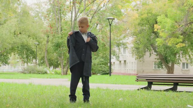 White boy preparing jacket in park near bench, adjusting sleeves and buttons with calm anticipation, morning light, trees and grass, student preparing to go back to school, candid outdoor portrait.