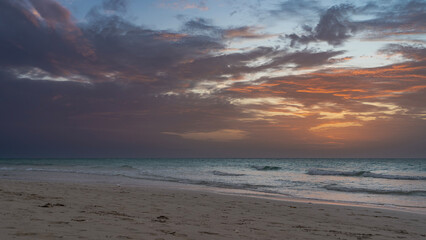 Obraz premium Tropical island, evening after sunset. The waves of the turquoise ocean roll towards the sandy beach. The purple clouds in the sky are highlighted in scarlet and gold. Cuba. Varadero.