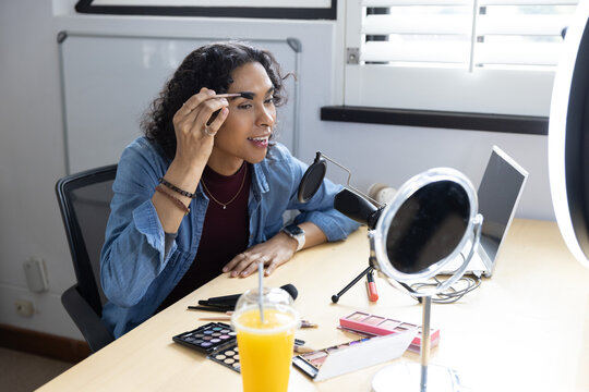 Non-binary adult applying eyebrow makeup at home studio desk, using magnifying mirror and mic
