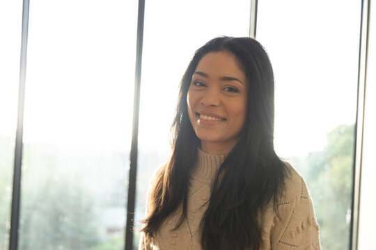 Smiling woman standing in front of glass panels in cream knit sweater, sunlight rim-lighting hair