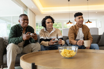 Three male friends playing console games on gray sofa at home holding controllers and chips © wavebreak3
