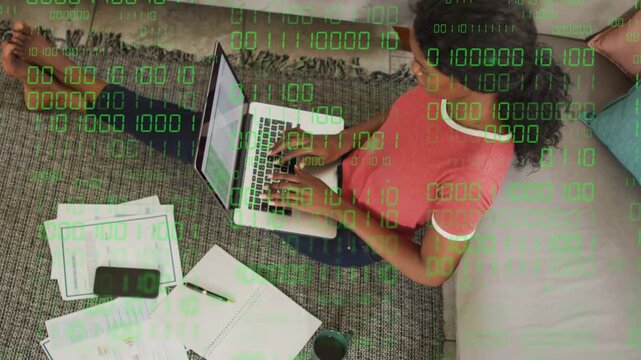 Woman typing on laptop on rug, activating green binary overlay while working remotely with papers