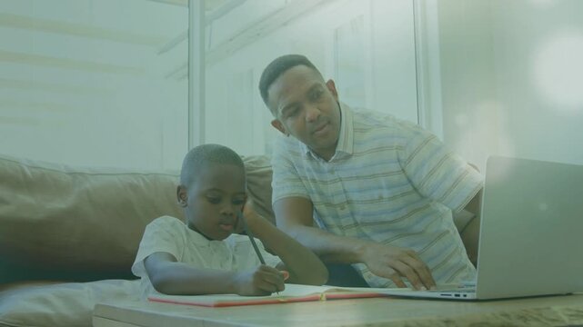 Father reaching for laptop, leaning toward notebook and guiding son writing homework for education