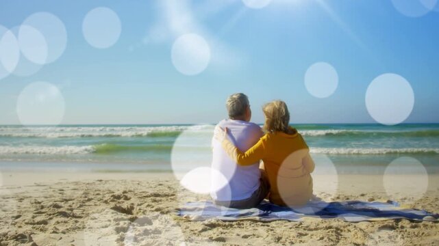 Woman turning draping arm around man on blanket watching waves, bokeh drifting over couple, travel
